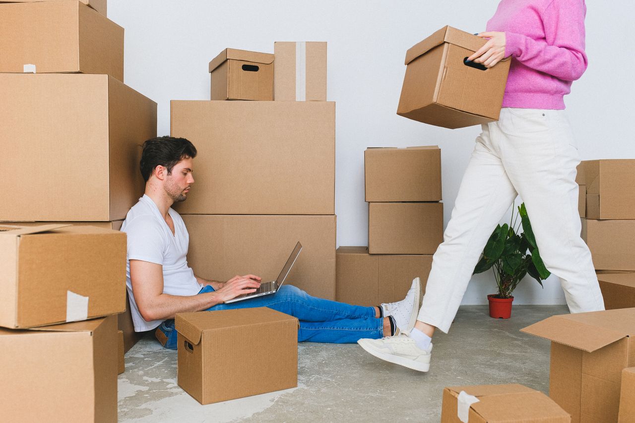 Side view of concentrated young male freelancer in casual clothes working remotely on laptop near crop anonymous girlfriend carrying cardboard boxes during relocation into new flat