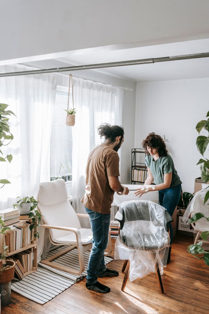 A couple efficiently packs furniture in a bright apartment with natural light and plants.